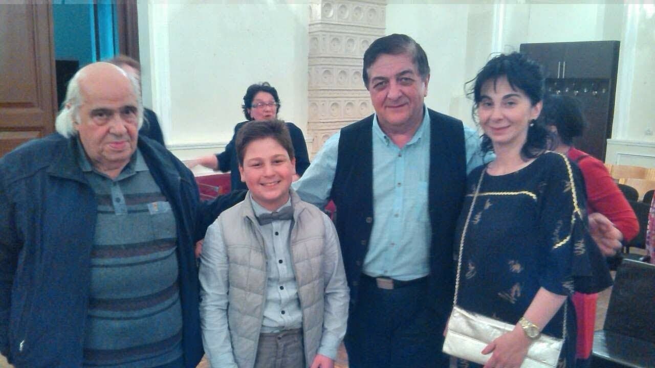 Aleksandre as a boy in a bow tie, smiling with a group of adults after a performance.