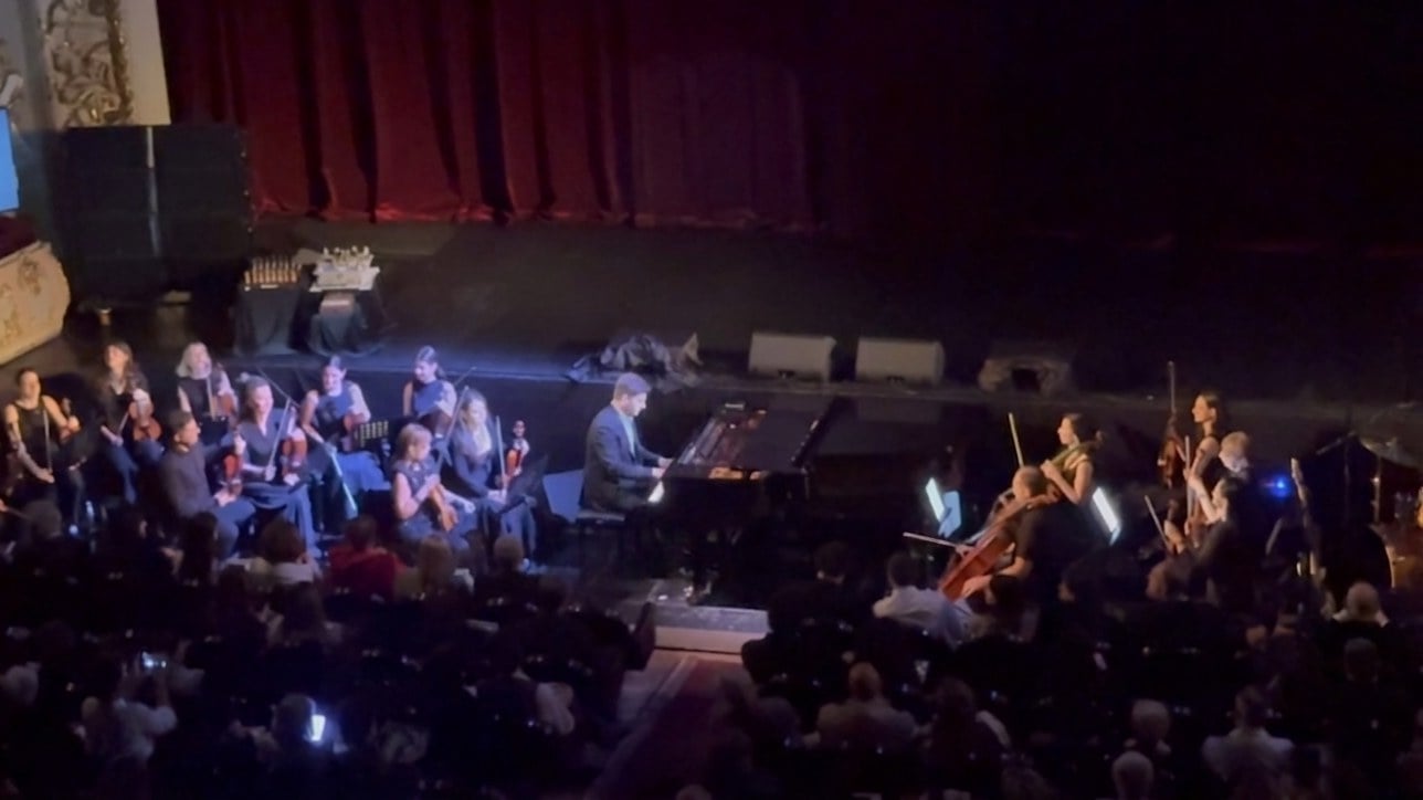 A pianist at a grand piano on a theatre stage, surrounded by a string orchestra, seen from the balcony.