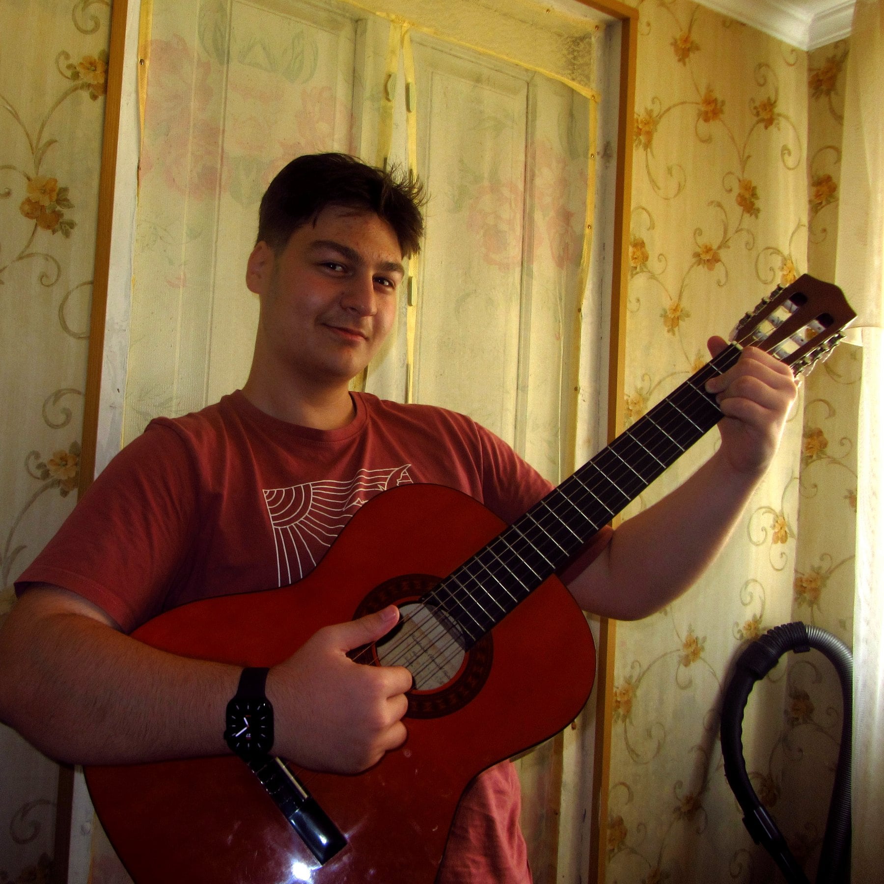 Aleksandre smiling at the camera while holding a classical guitar at home.